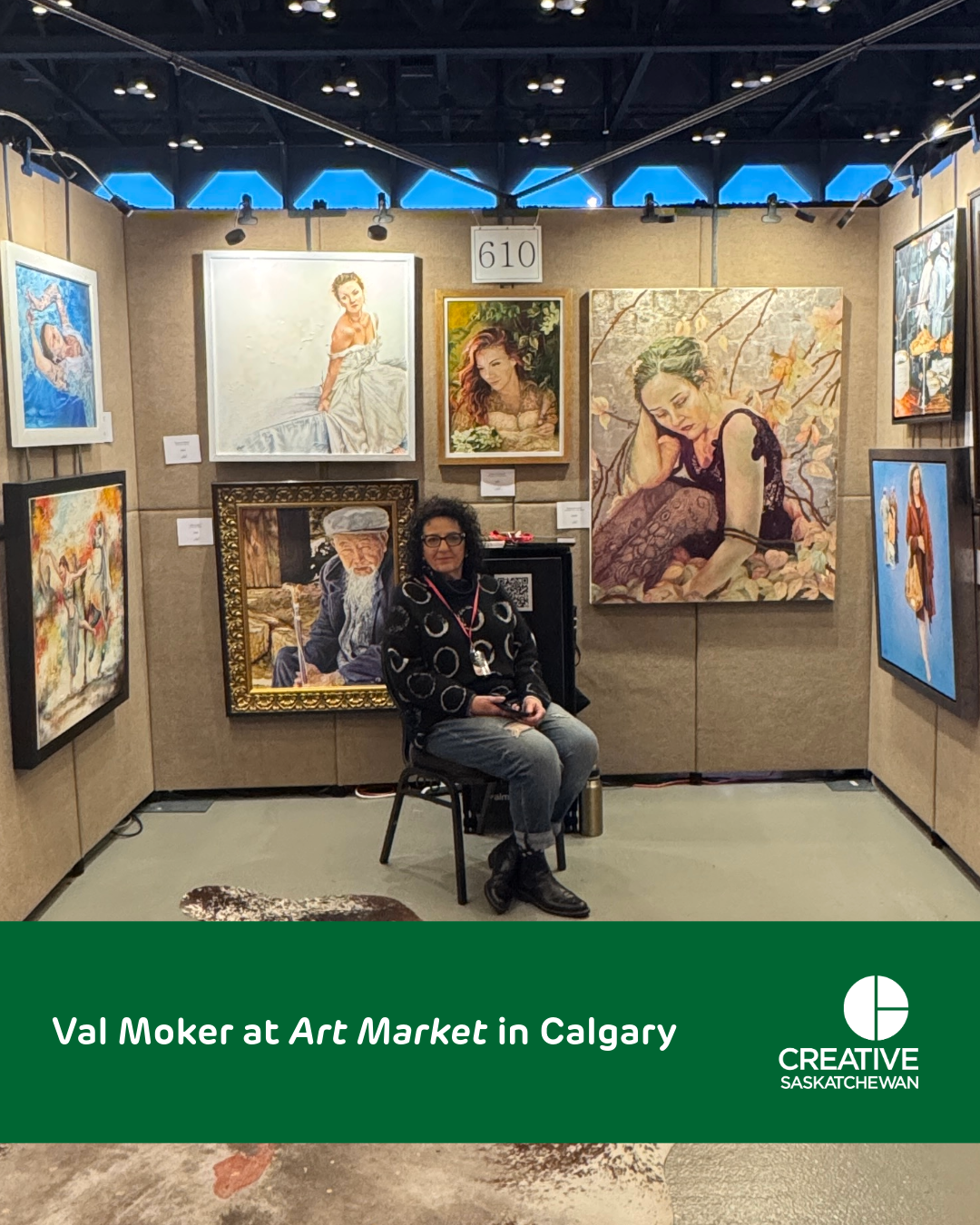 Val sits on a chair in a gallery booth, her artwork surrounds her on the walls.
