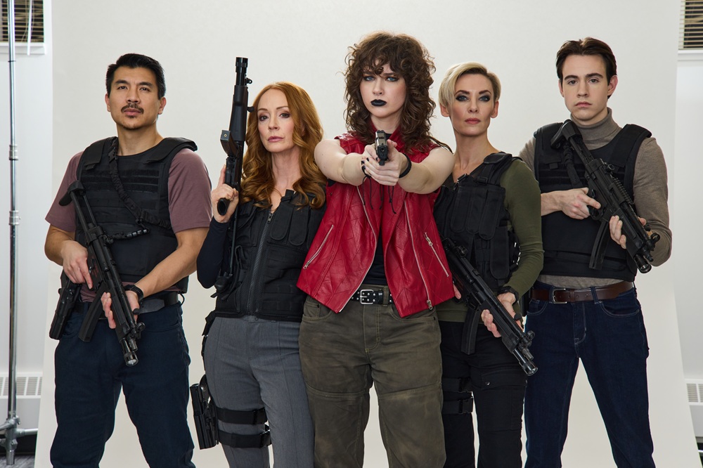 A group of two men and three women in combat gear hold guns and stare down the camera with an attitude. The woman in the centre stands out in a red vest with black lipstick and curly, dark hair.