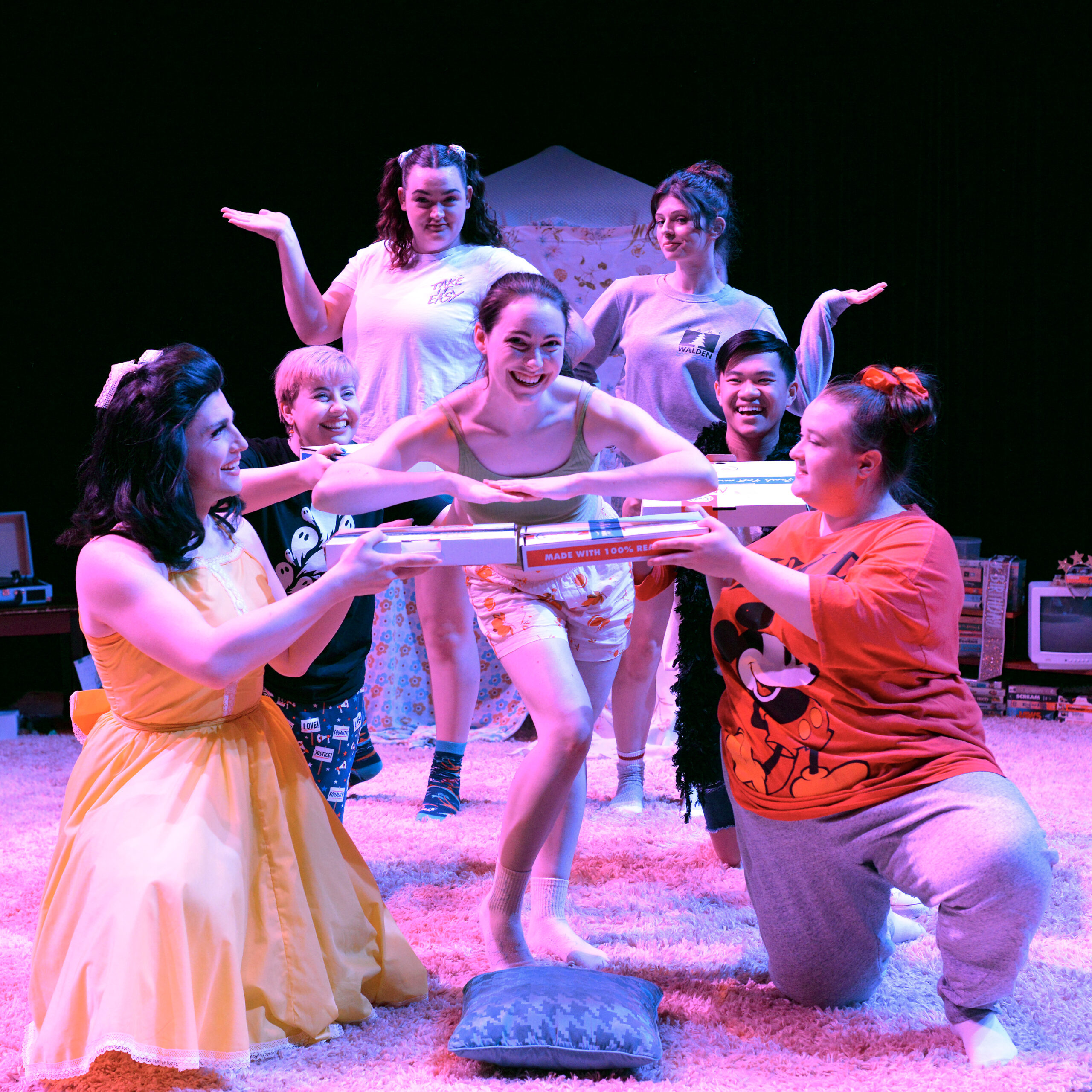 Actors posing on stage, all of them are wearing pajamas except one who wears a yellow dress. Four of them hold pizza boxes around a girl who poses in the center. 