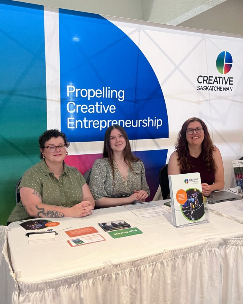 Three smiling women sit behind a table displaying brochures and hand outs for Creative Saskatchewan. There is a colourful Creative Saskatchewan backdrop behind them.