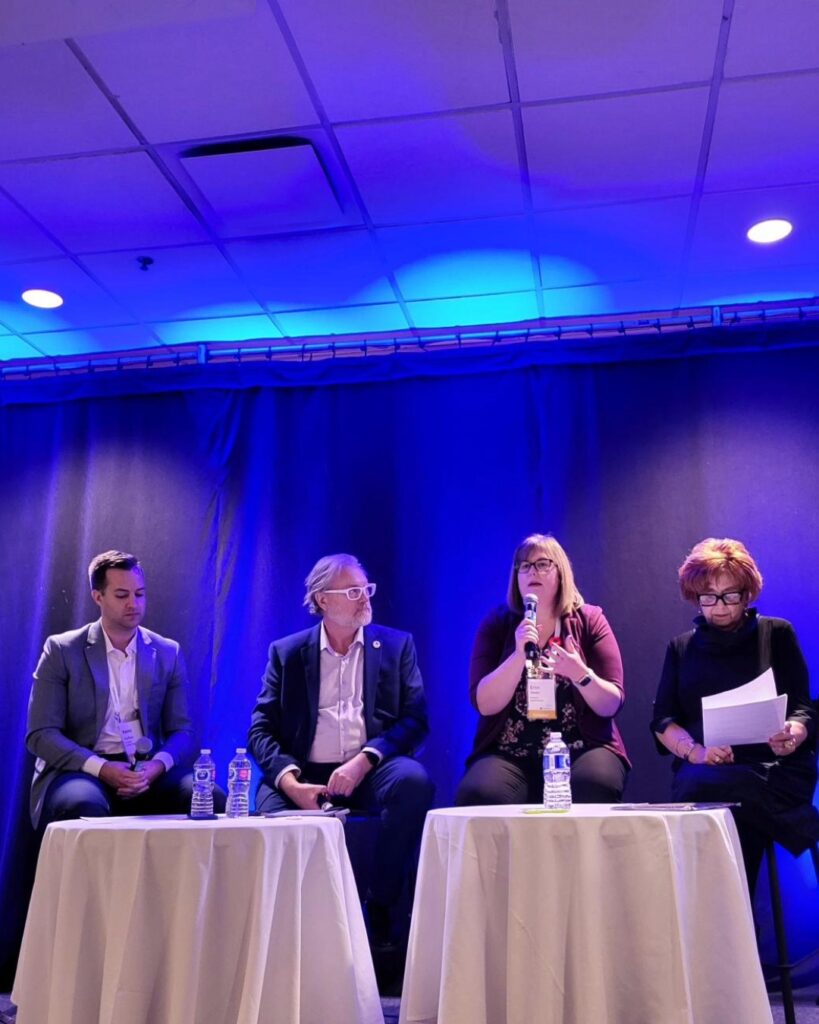 Four panelists sit on stools on stage. Each have a microphone, and one of them is speaking while another checks their notes.
