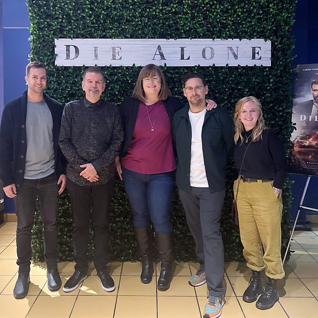 A group of people pose smiling in front of a plant backdrop with a "Die Alone" banner. A movie poster for the movie "Die Alone" is beside them.