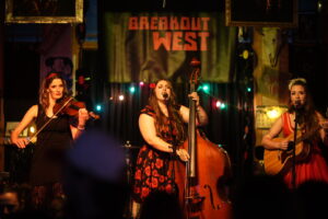Two women singing into microphones on stage, a black banner hangs behind them that says “BREAKOUT WEST” in red block letters. The woman on the left is playing a stand up bass, she has waist-length brown hair with a red rose tucked behind her ear, and is wearing a black dress patterned with red roses. The woman on the left plays an acoustic guitar, she has waist-length brown hair with a white feather hairpiece on top of her head. She wears a red dress with white trim. A purple bar across the bottom of the image reads “Gil & Wil perform at BreakOut West” 