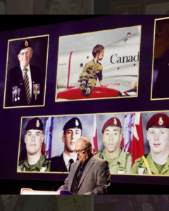 A man wearing a grey suit and black wire-frame glasses stands behind a large black podium, head bowed as he speaks into the microphone. Behind him are large photos of various veterans, young and old, and in the centre is a painting of a young boy dressed in military uniform sitting on the wing of a Canadian war plane. 