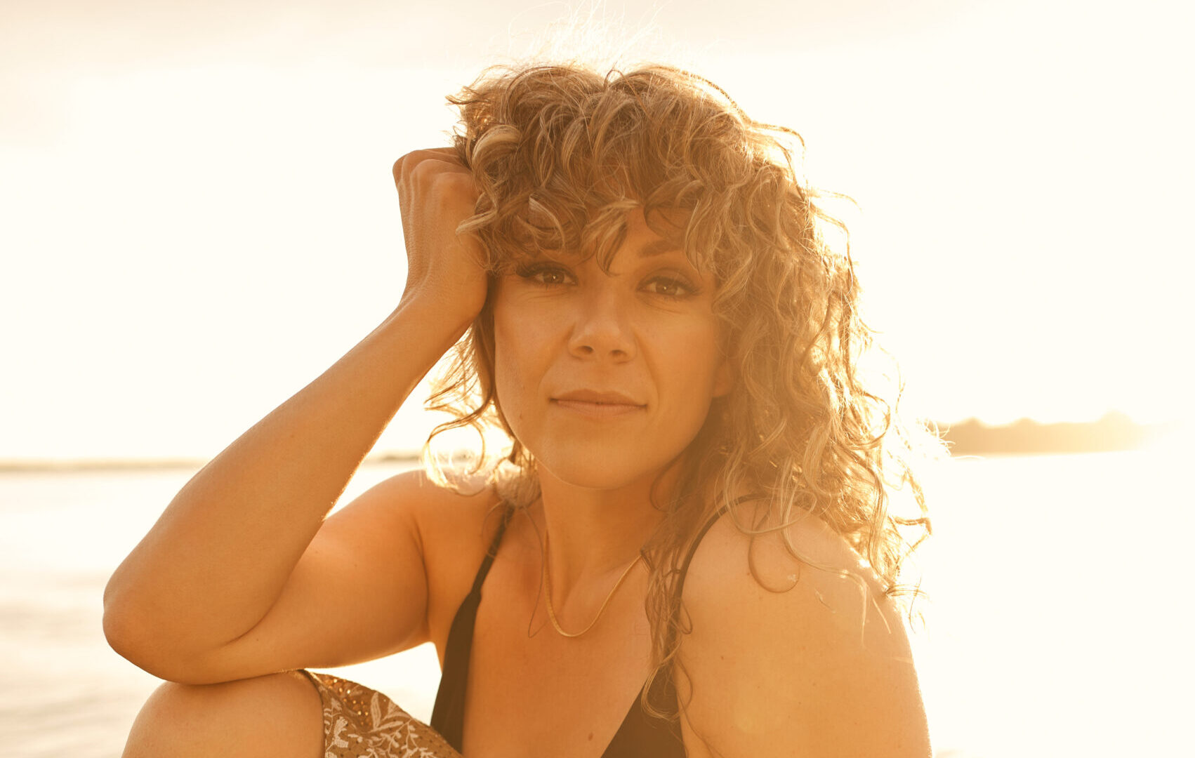 A woman with curly blonde hair lounges casually on a dock, bathed in golden sunlight. She wears a black tank top and a patterned skirt.