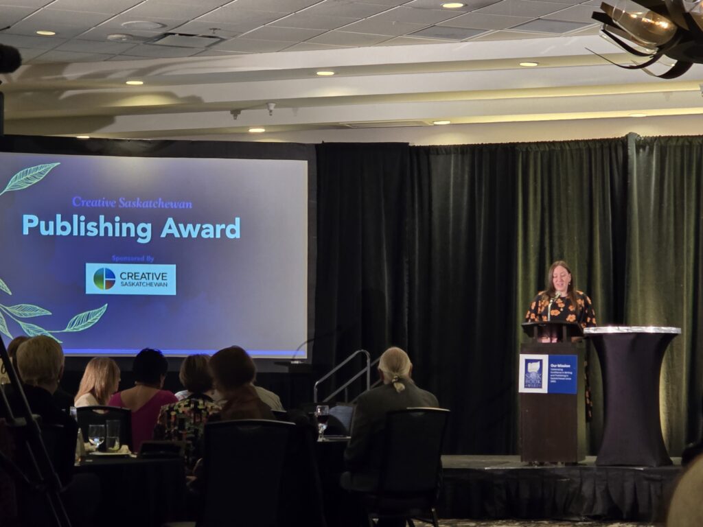 A woman with dark and wearing a flowery dress stands at a podium to present an award. A screen beside her reads "Publishing Award" with the Creative Saskatchewan logo.
