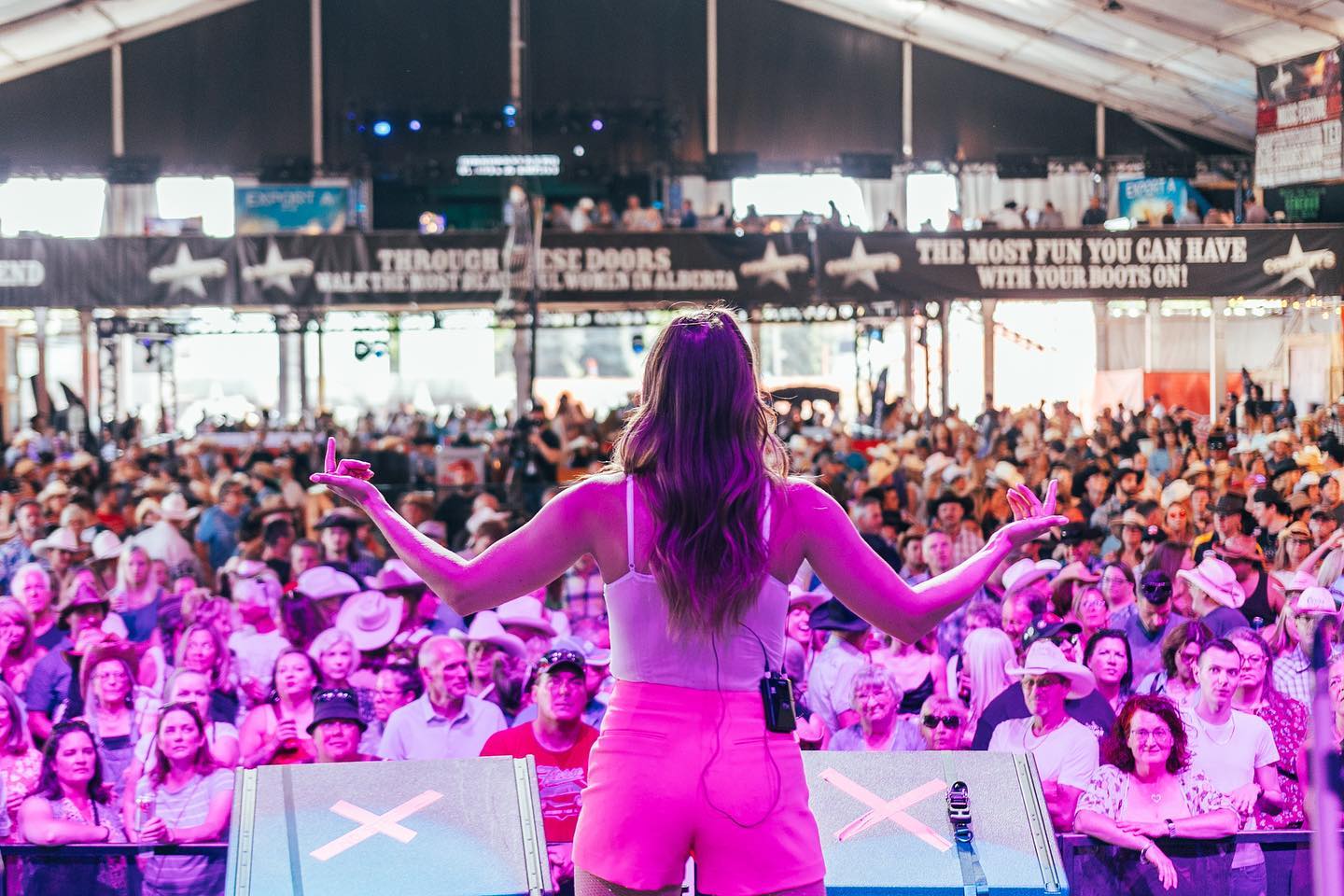 A female country music singer has her back to the camera as she stands on stage facing a huge crowd. She is bathed in pink light and is wearing pink shorts and a white tank top.