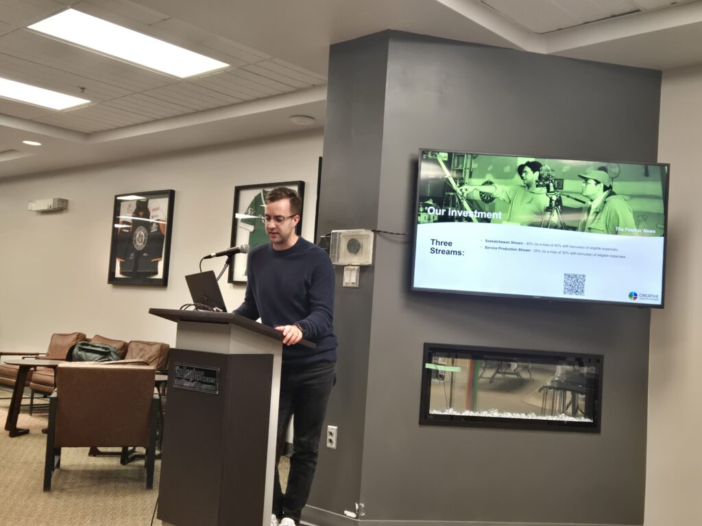 A man with brown hair and wearing a blue sweater and glasses is presenting at a podium. A TV screen behind him shows a slide containing film industry data.